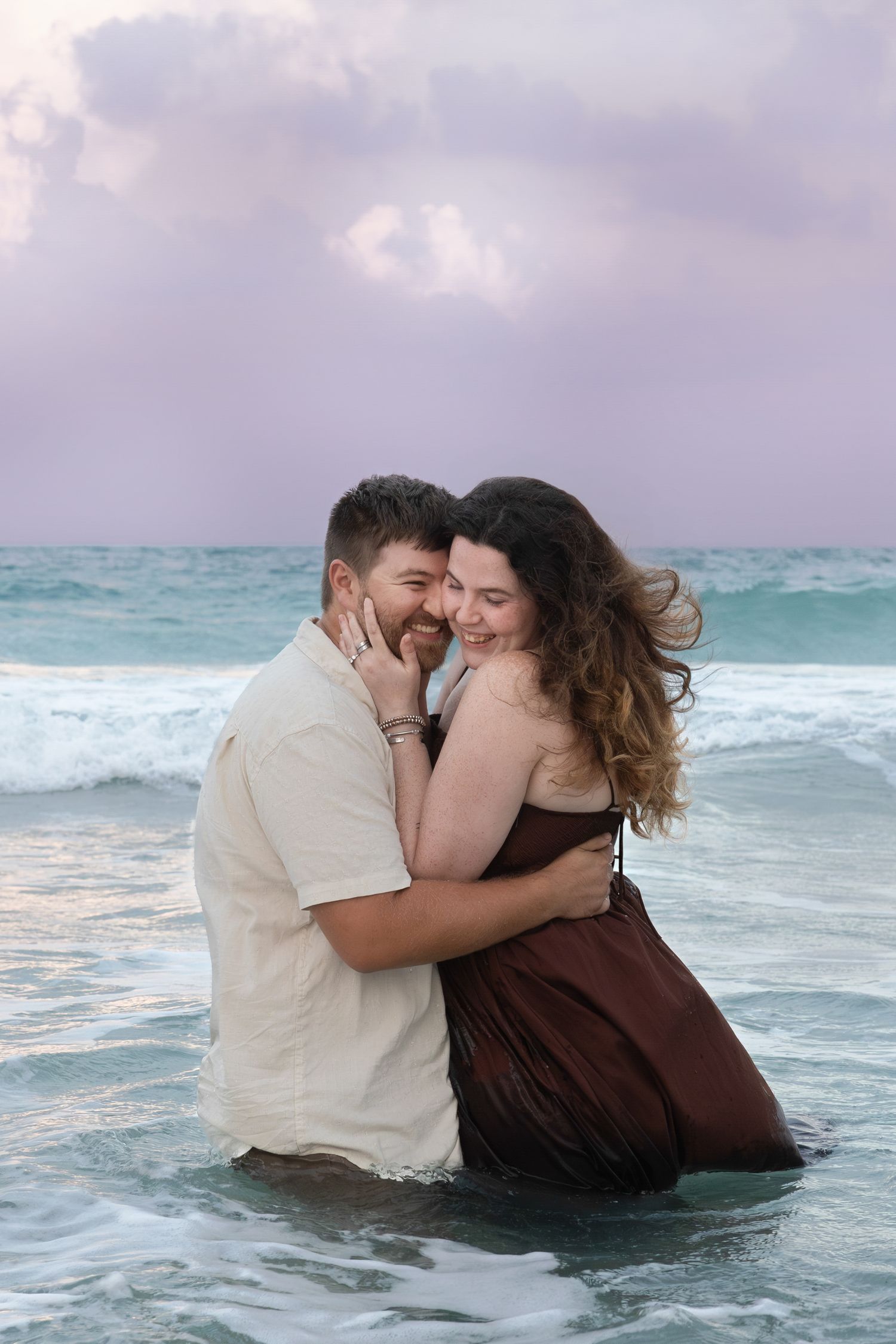 Happy couple embracing and smiling in ocean waves at sunset on the beach.