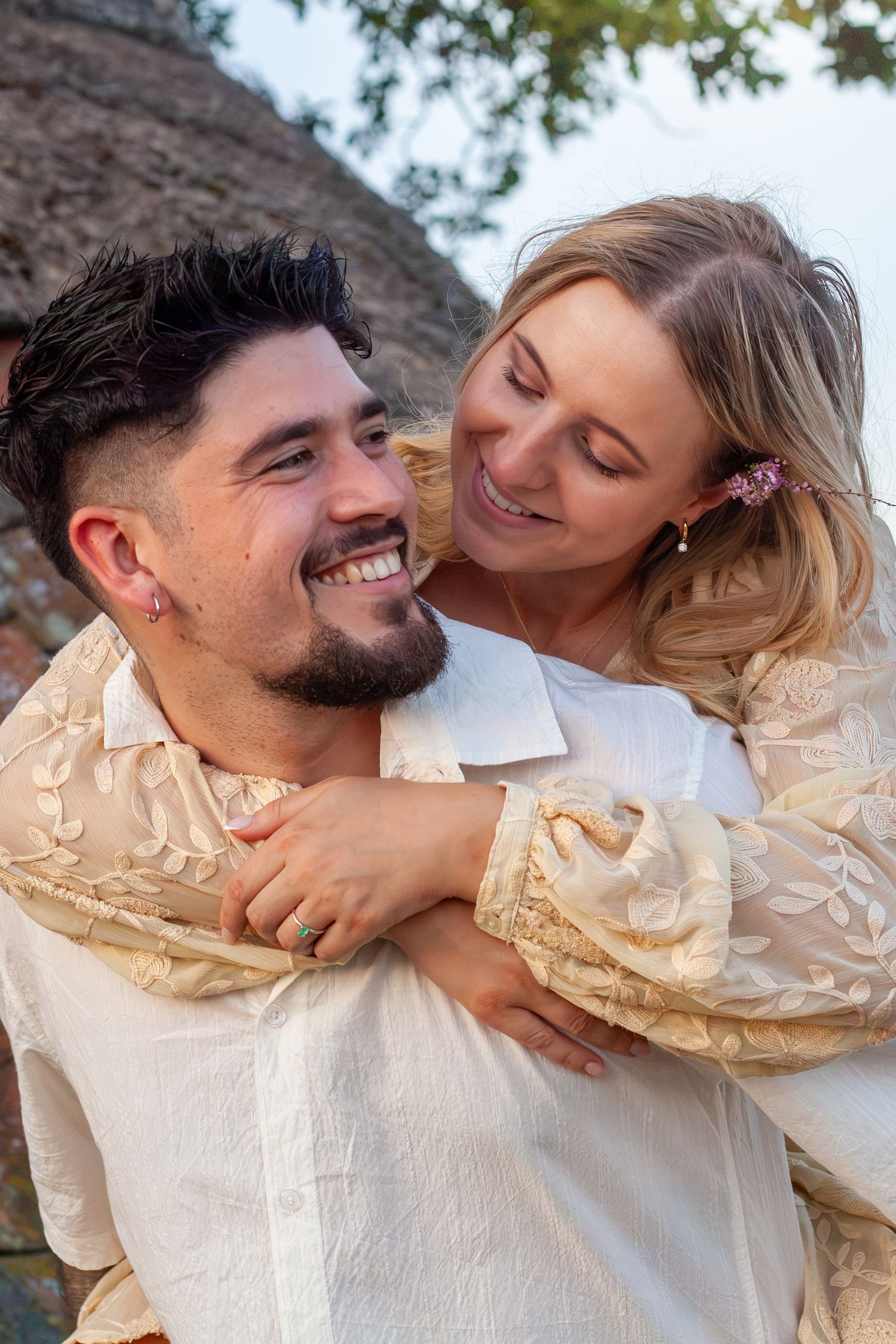 A smiling couple in matching cream outfits share a joyful piggyback moment outdoors, with the woman hugging the man from behind.