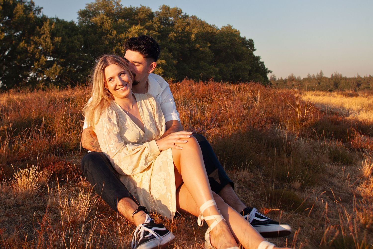 Happy couple sitting in golden field at sunset, woman in floral dress smiling as man embraces her from behind.