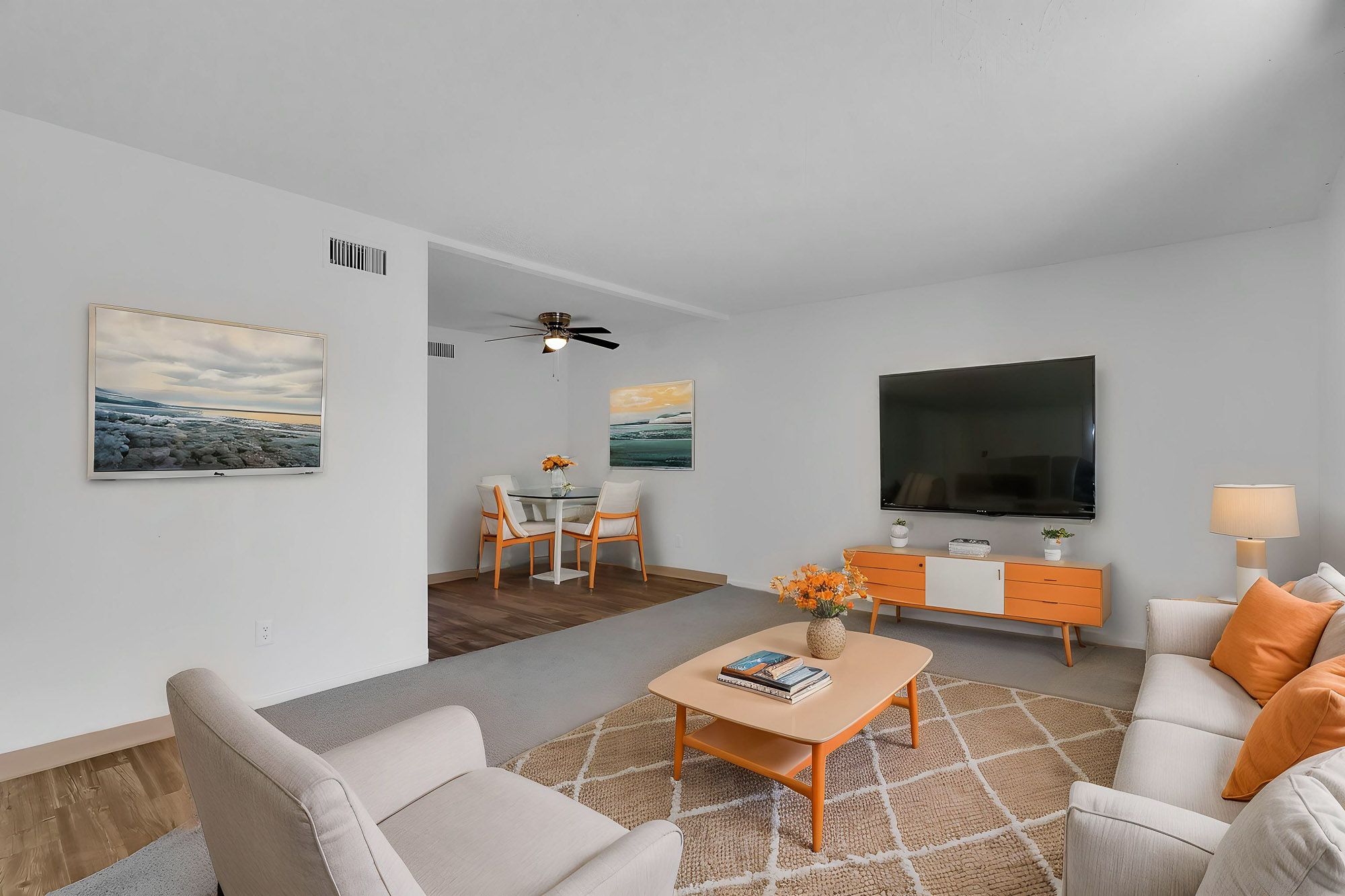 Bright living room with neutral sofas, ceiling fan, orange accents, wall art, and open dining area beyond.