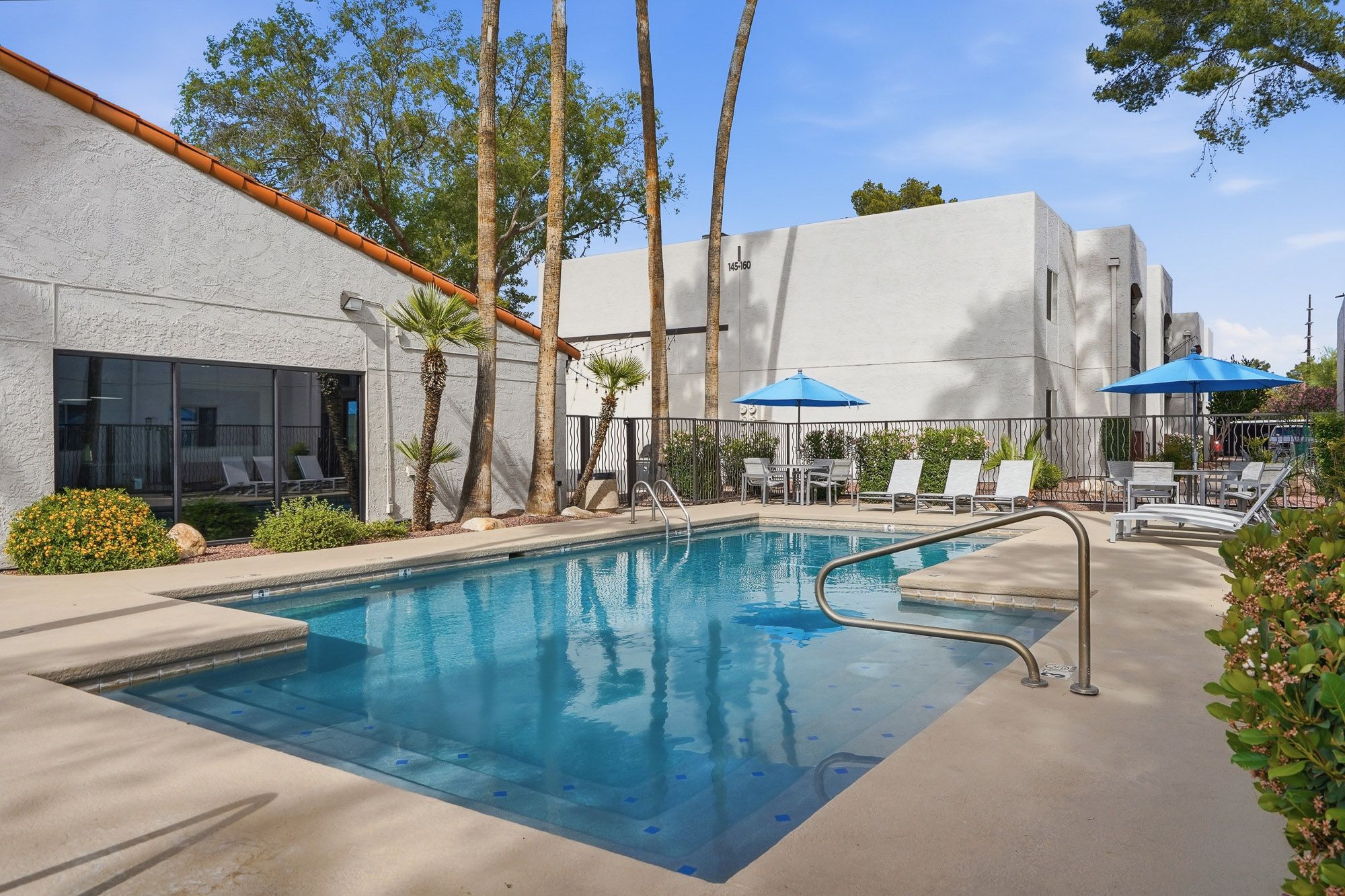 Sparkling outdoor pool surrounded by lounge chairs and blue umbrellas at a sunny apartment community with palm trees.