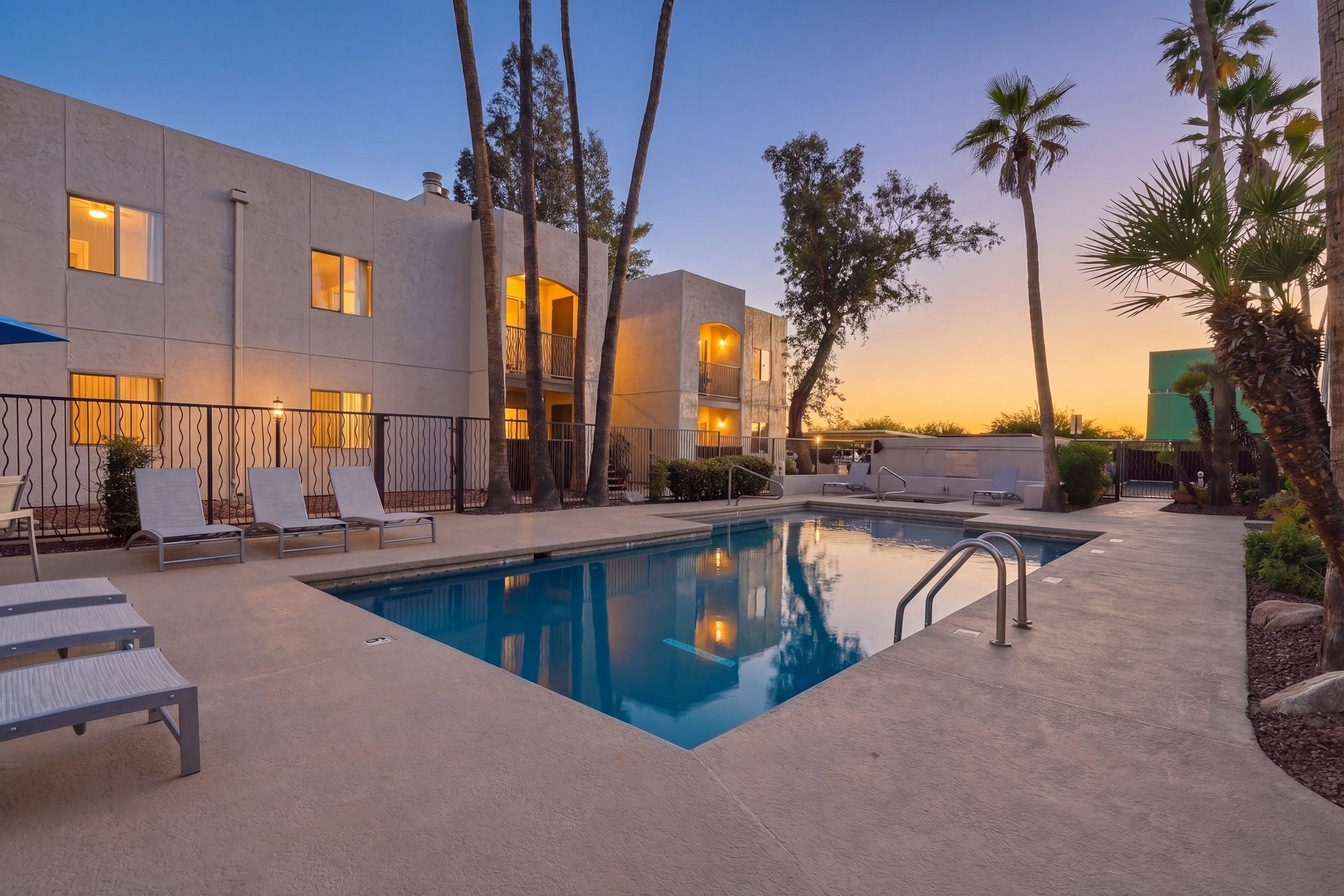 Resort-style outdoor pool with lounge chairs at dusk, surrounded by palm trees and apartment buildings.