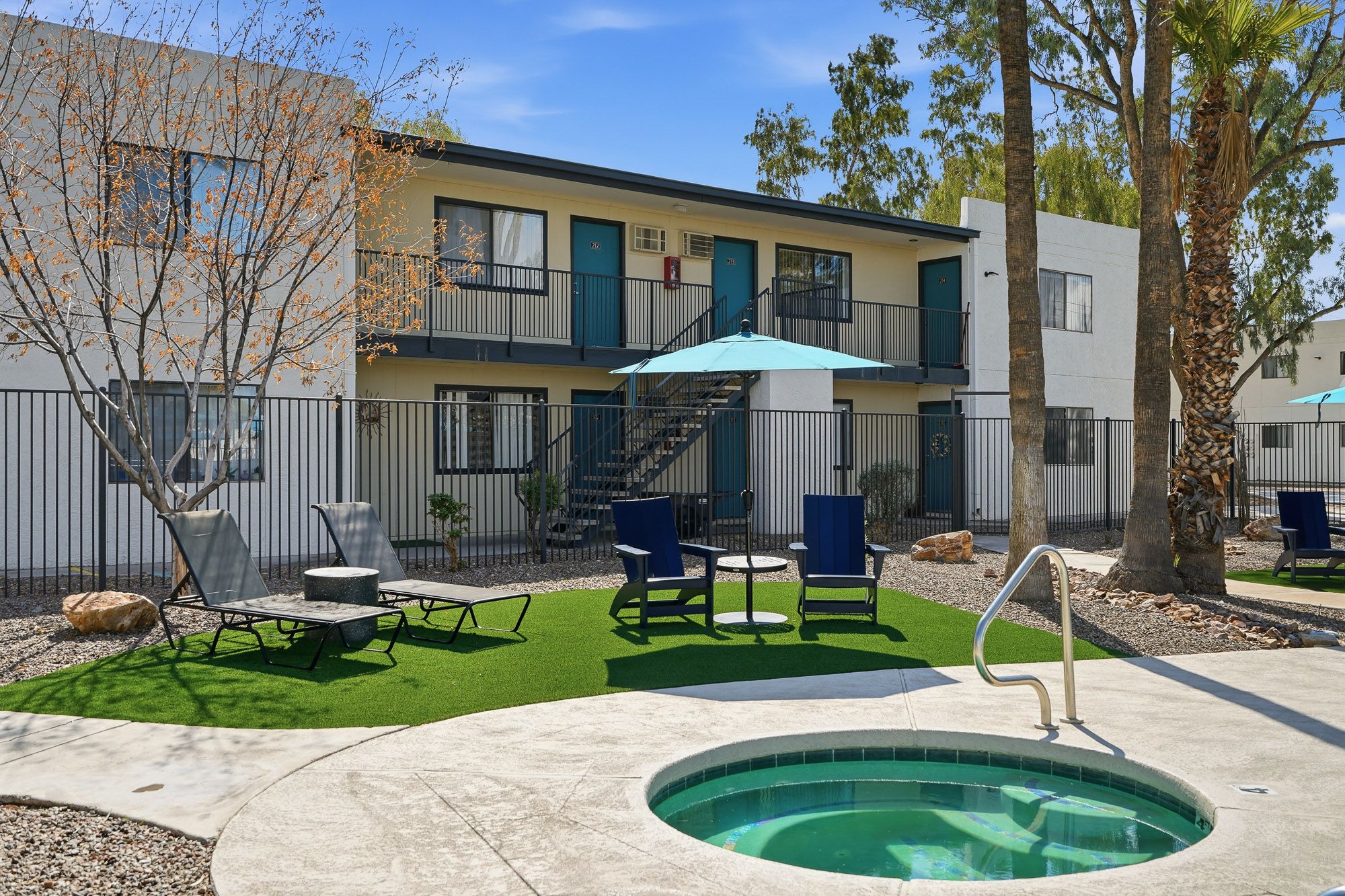 Outdoor hot tub and patio seating on artificial turf beside a two-story white stucco apartment building.