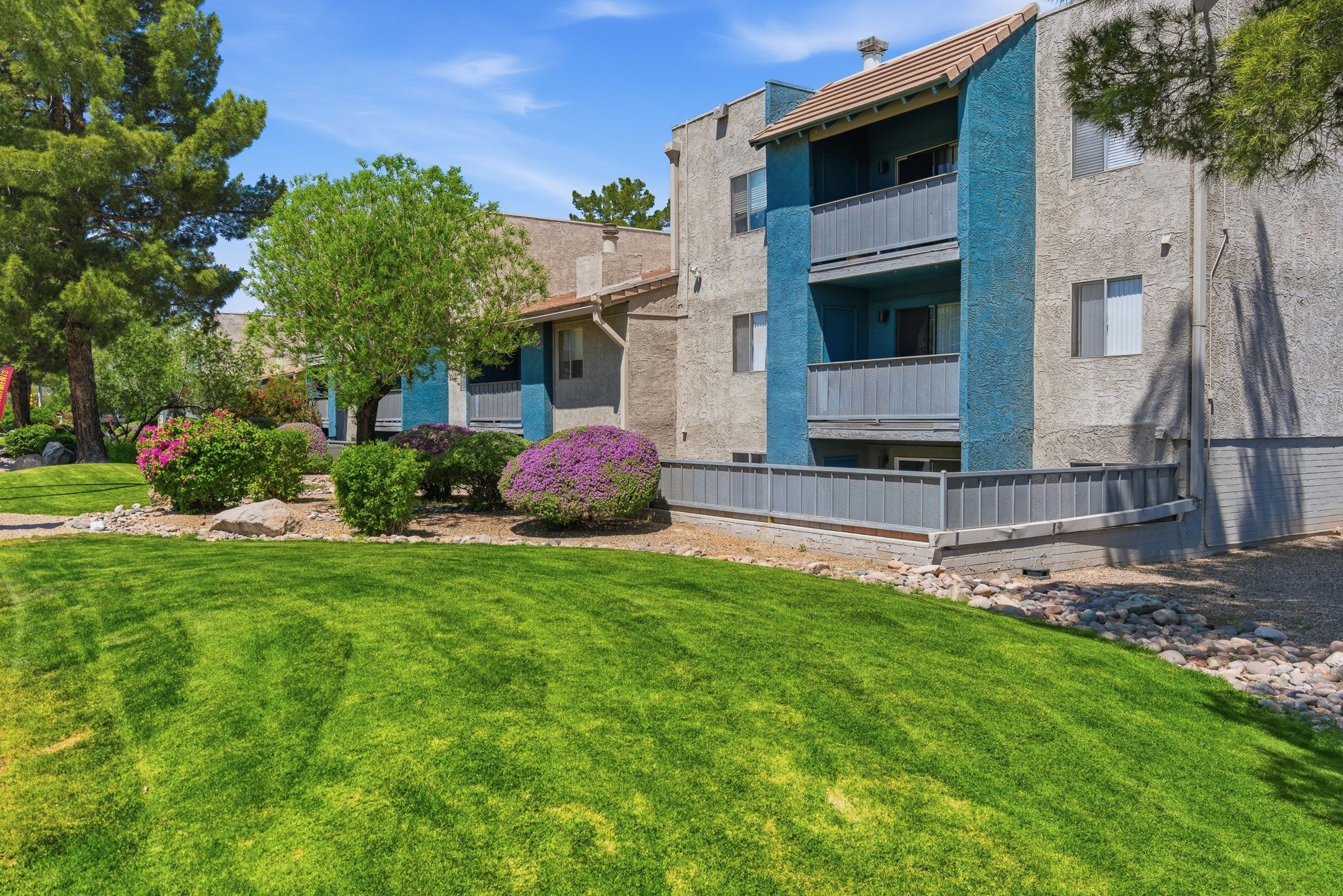 Lush green lawn surrounds a two-story apartment complex with teal balconies and colorful landscaping under a bright blue sky.