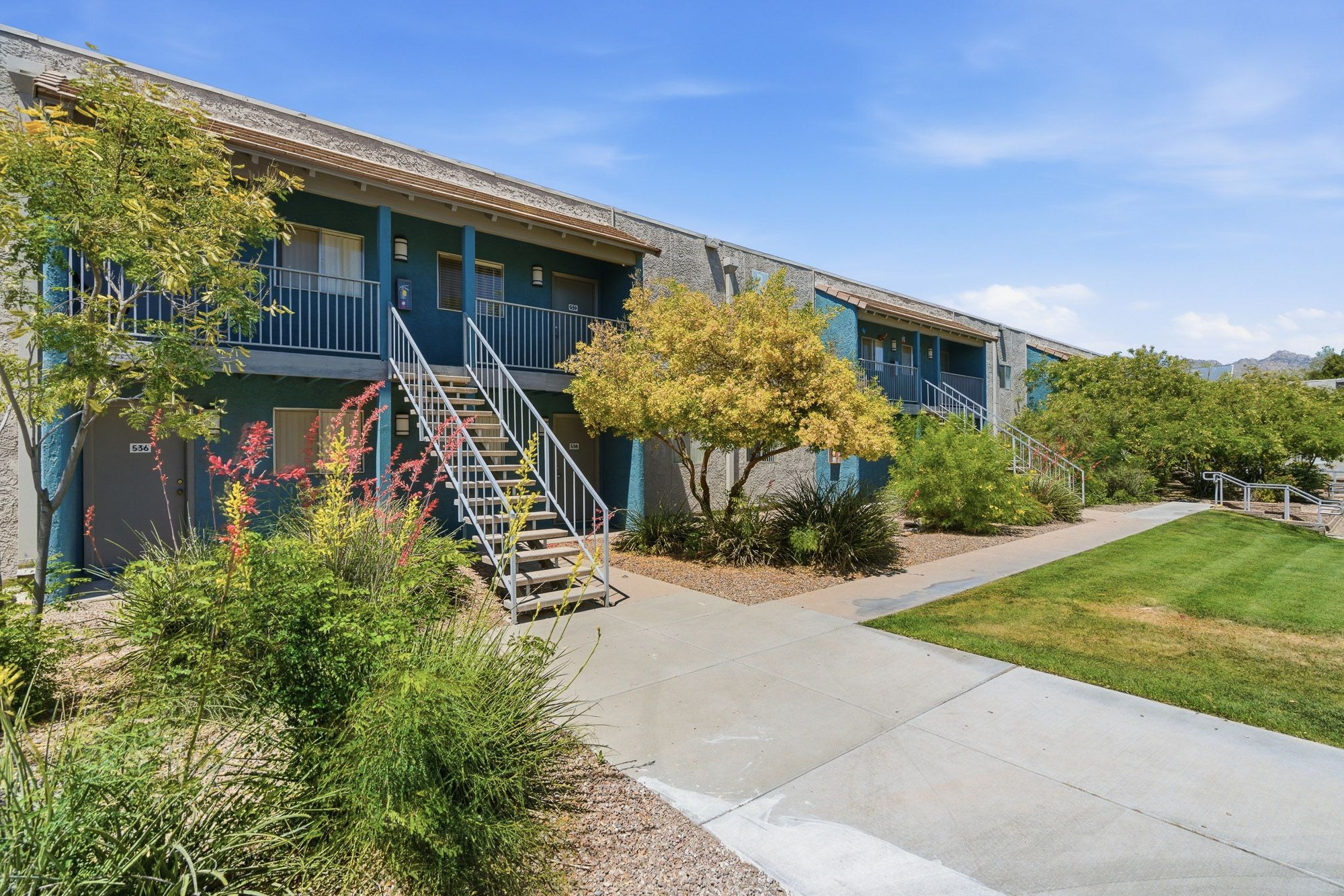 Blue two-story apartment complex with lush landscaping, staircase, and walkway under a clear sky.