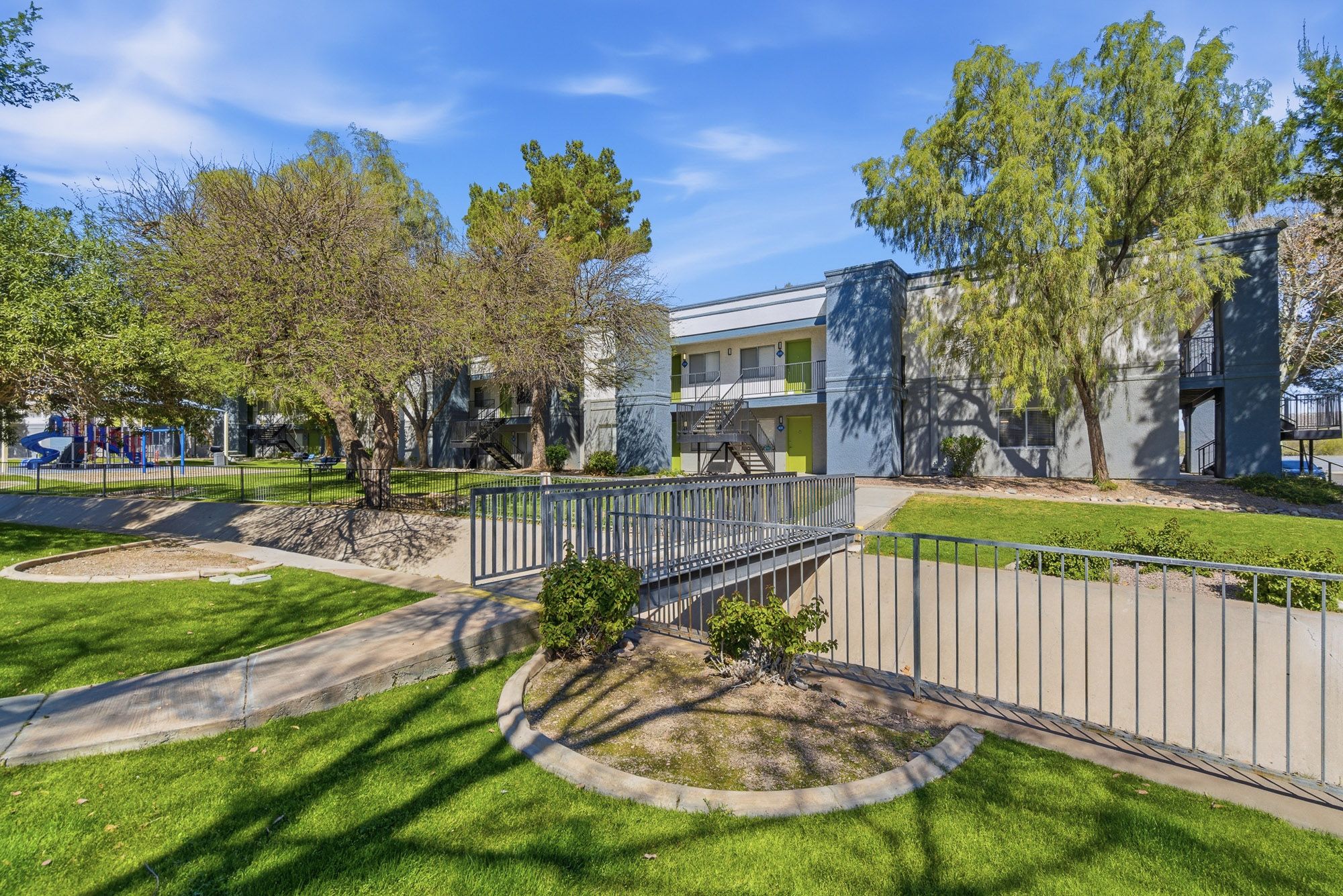 Apartment complex exterior with manicured green lawn, walking path, and leafy trees on a sunny day.