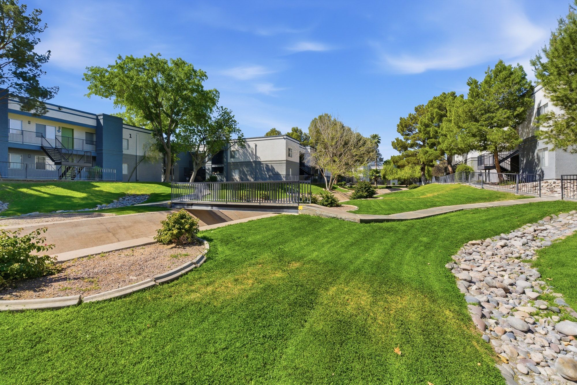 Lush green lawn with landscaped gardens and mature trees surrounding a residential apartment complex on a sunny day.
