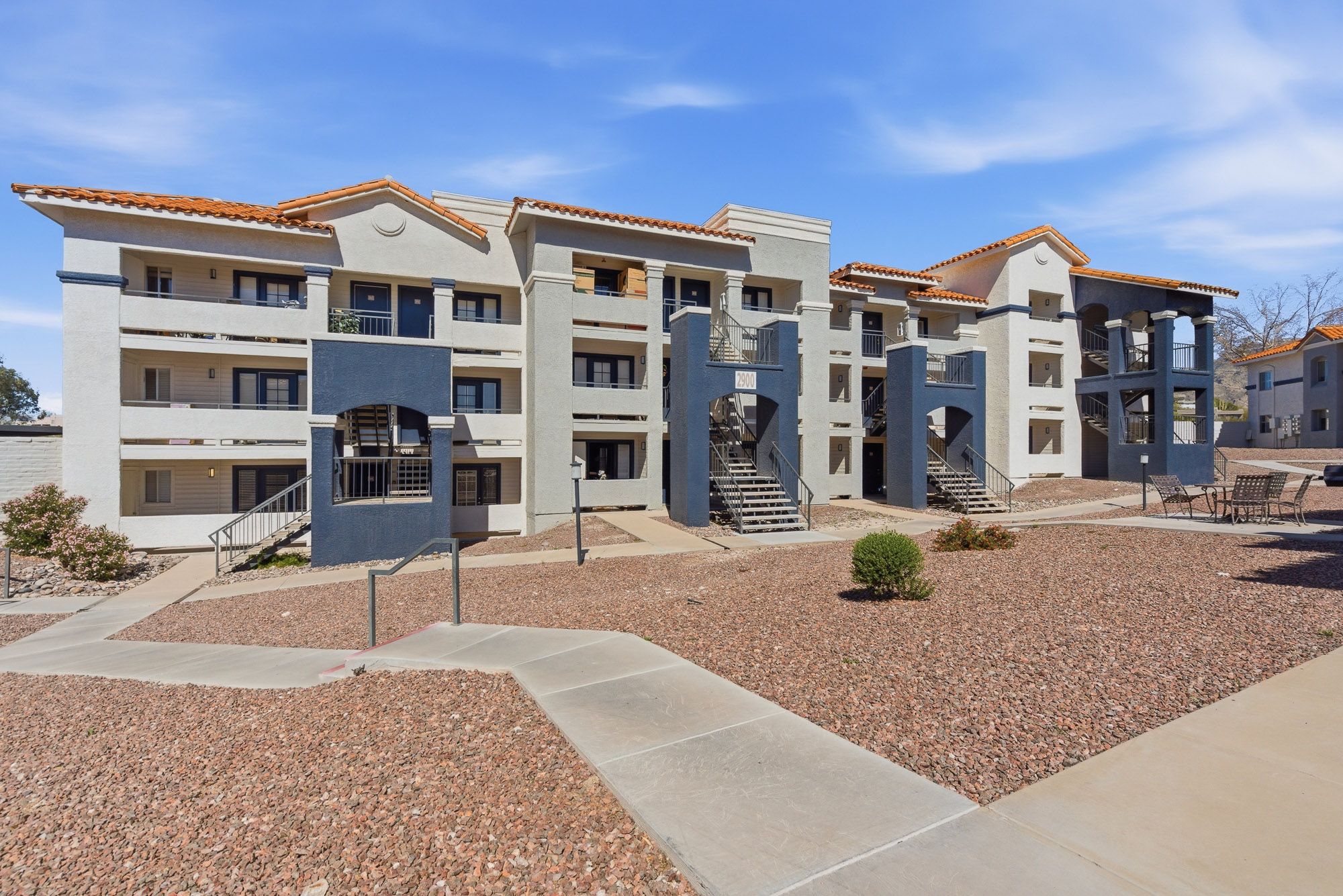 Three-story apartment complex with desert landscaping, balconies, and neutral stucco exterior under a clear blue sky.