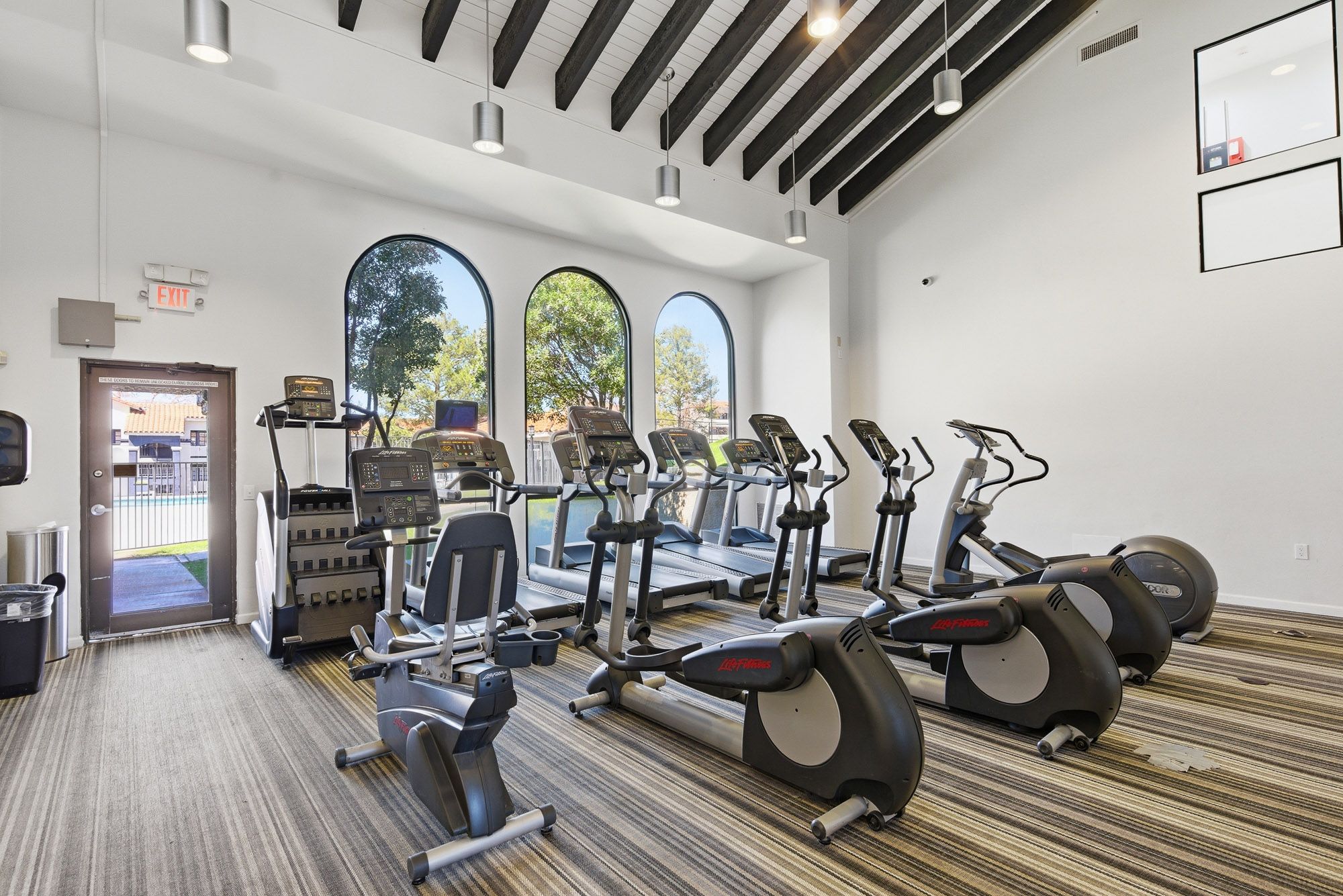 Modern apartment fitness center with cardio machines, arched windows, and striped flooring under exposed beam ceiling.
