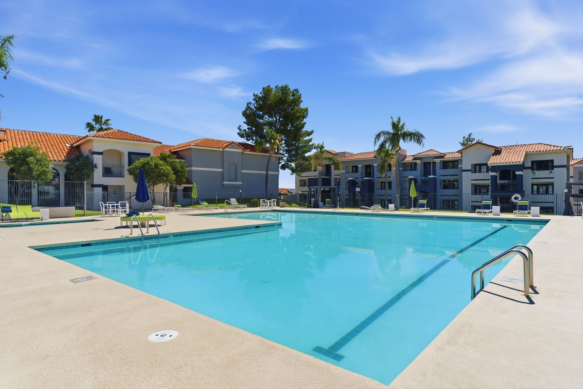 Sparkling outdoor pool surrounded by lounge chairs at a sunny apartment complex with palm trees and blue sky.