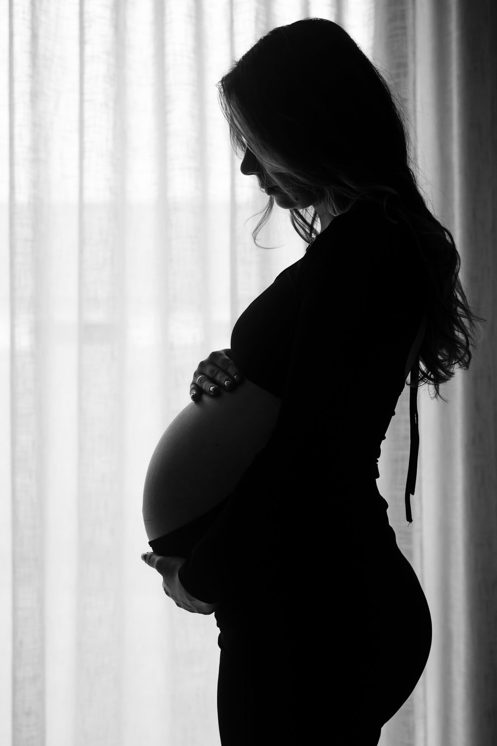 A silhouetted pregnant woman stands in profile by a window, tenderly holding her belly in black and white.