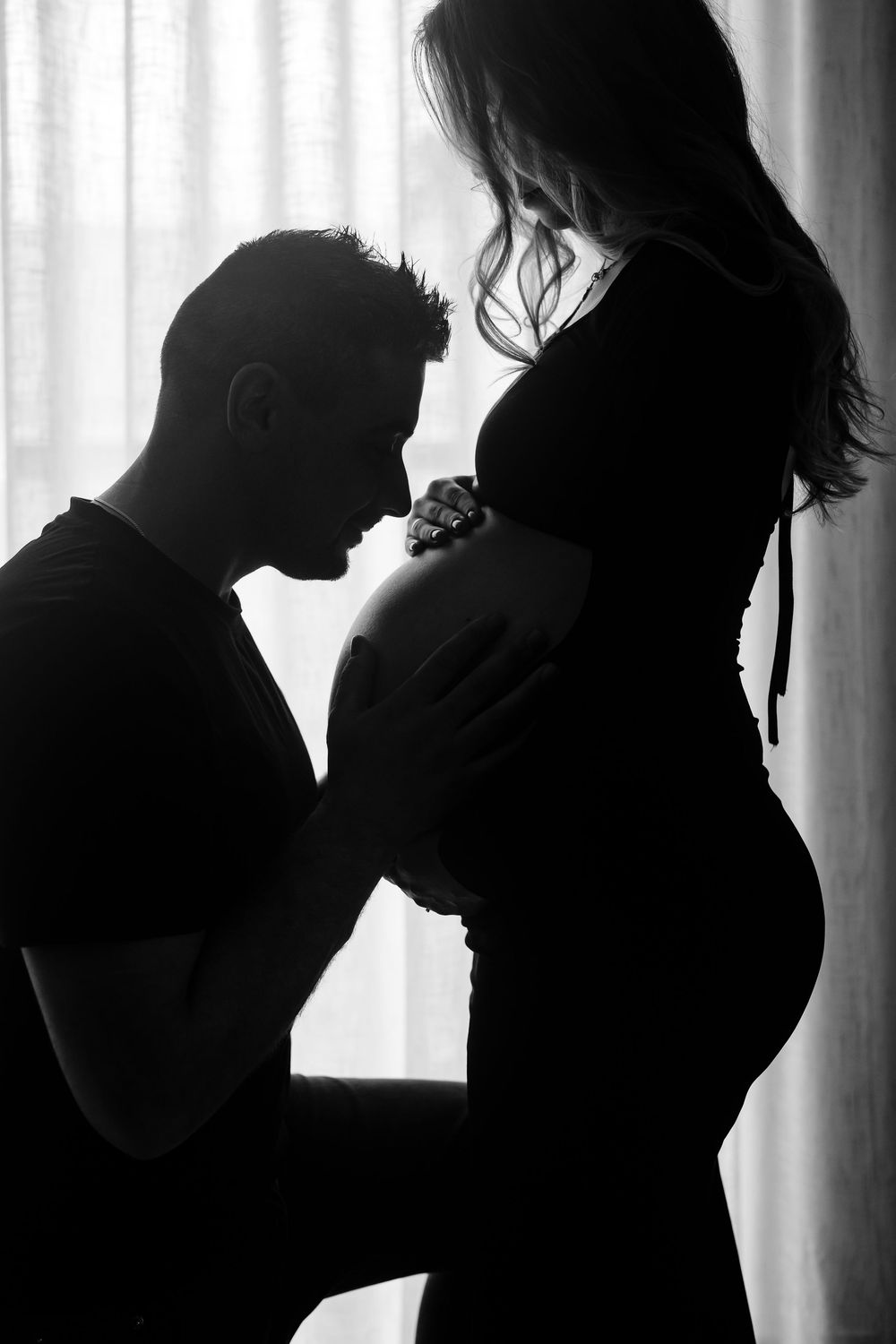 A series of silhouetted figures in romantic poses against a bright window backdrop creates an intimate maternity photoshoot.