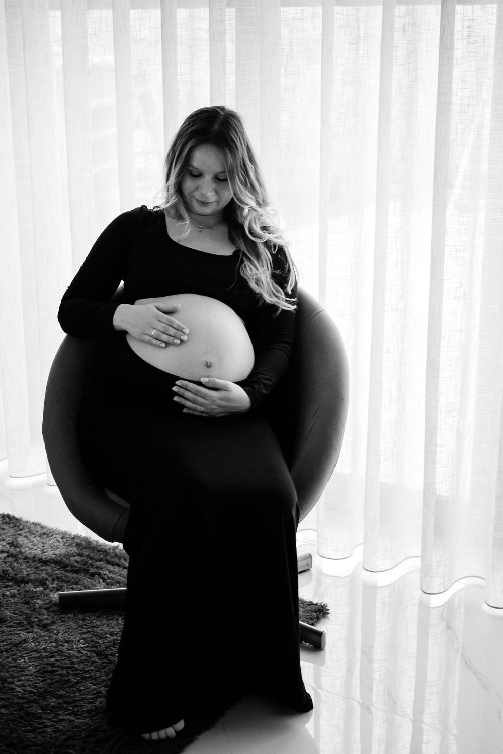 Black and white maternity photo series showing a pregnant woman in a dark dress touching her belly against white curtains.