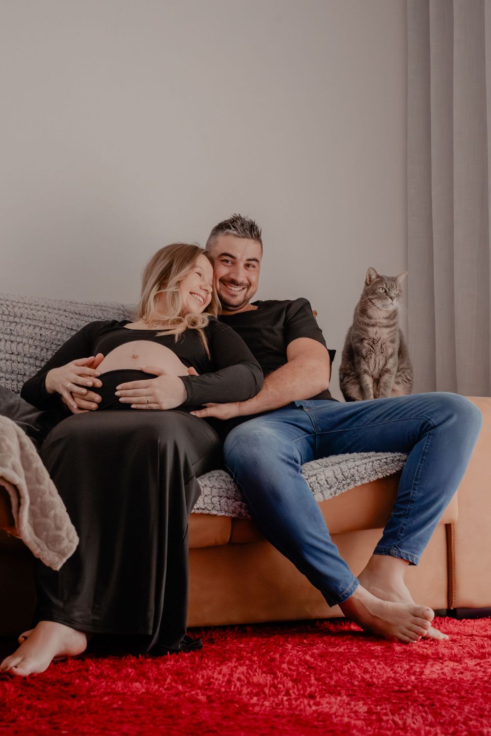 A couple relaxes on a gray couch with their gray cat, with a bright red rug beneath them.