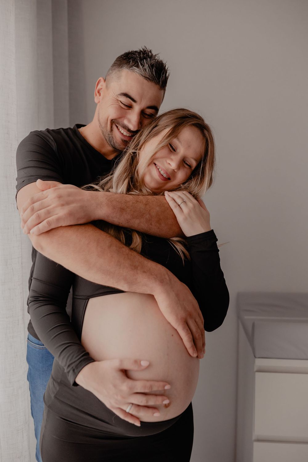 A couple embraces in a tender moment, showing affection and joy while wearing casual clothing in a bright indoor setting.