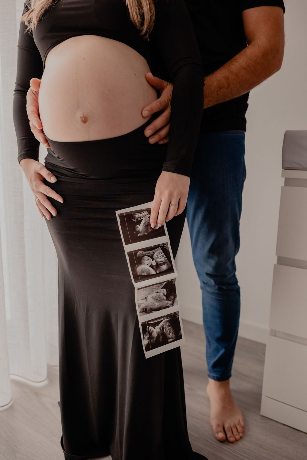 A pregnant woman in a black dress holds ultrasound photos while someone stands beside her showing her growing belly.