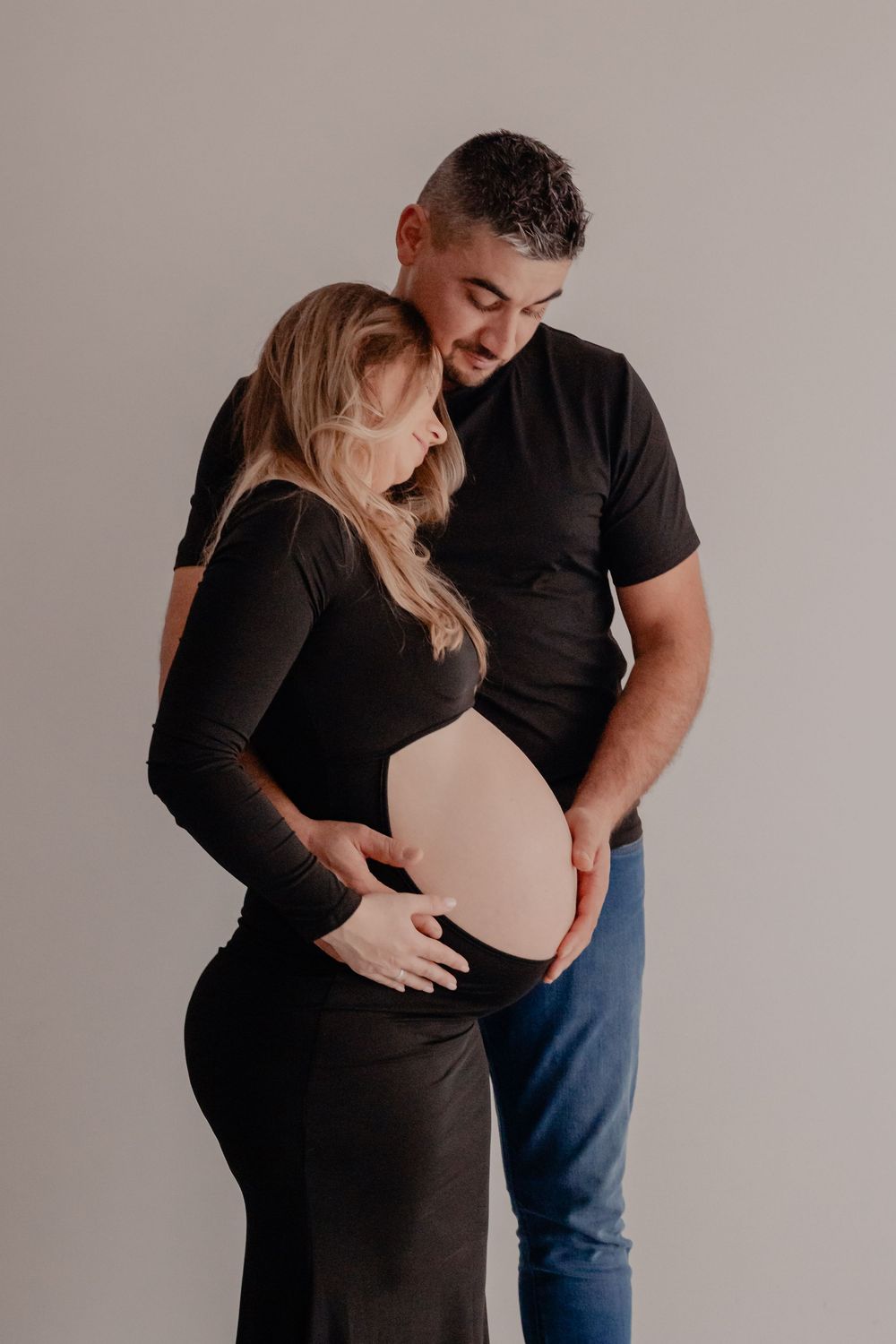 A couple in black clothing shares an intimate moment during a maternity photoshoot against a neutral background.