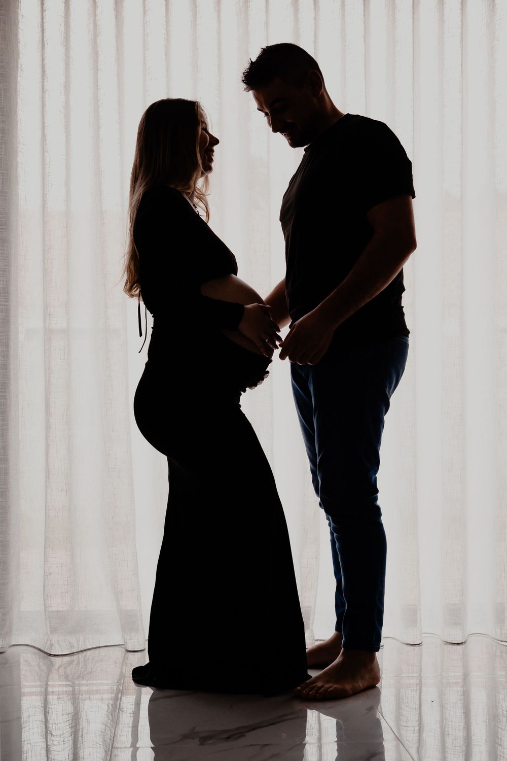 Silhouetted pregnant couple stands intimately by window in maternity photo shoot.