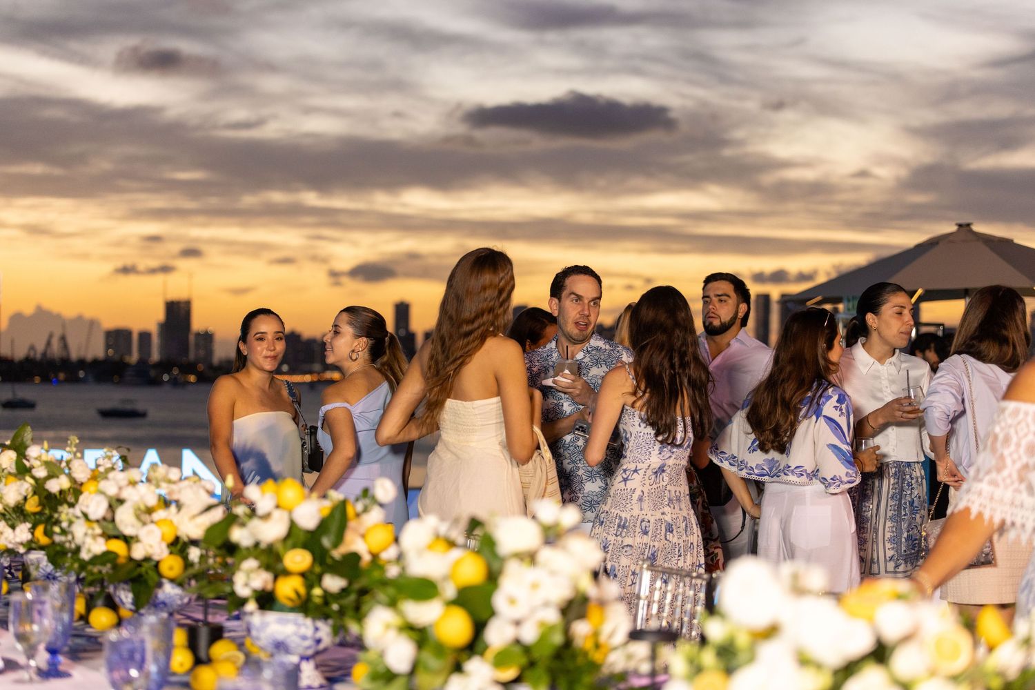 Guests mingle at an elegant outdoor waterfront wedding reception at sunset with floral arrangements.