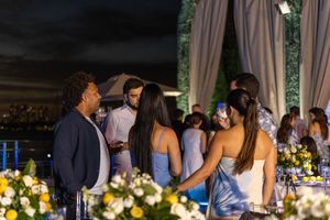 Elegant outdoor evening party with guests mingling near floral arrangements under draped canopy with city skyline backdrop.