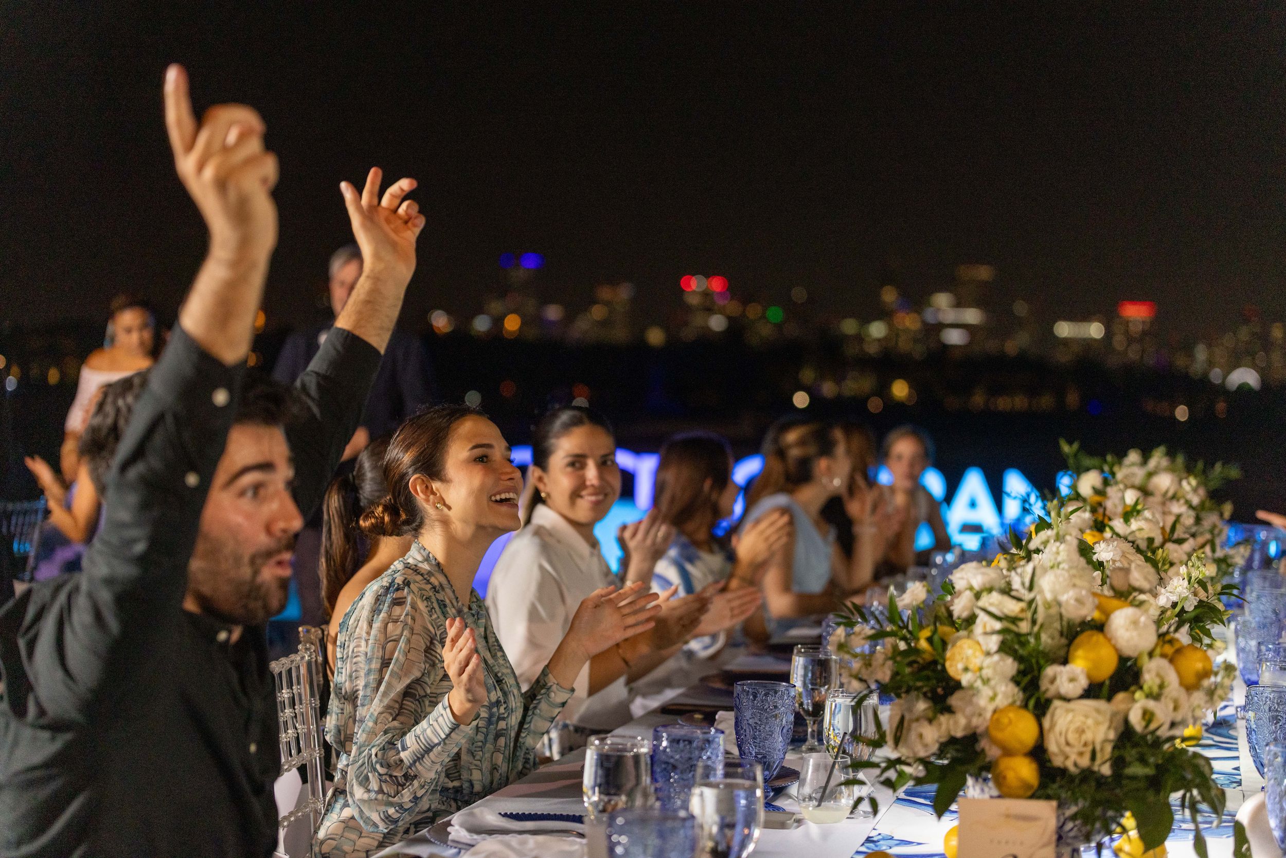 Guests clapping and celebrating at an elegant outdoor evening dinner party with city lights in the background.