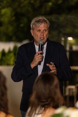 Middle-aged man in dark blazer speaking into microphone at outdoor evening event.