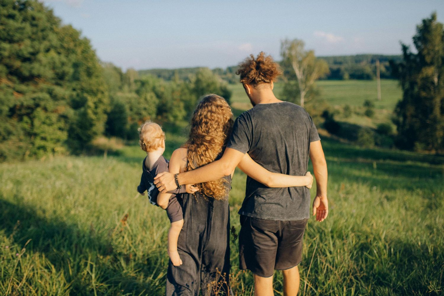 A family walks together through a sunlit meadow on a summer evening, viewed from behind.