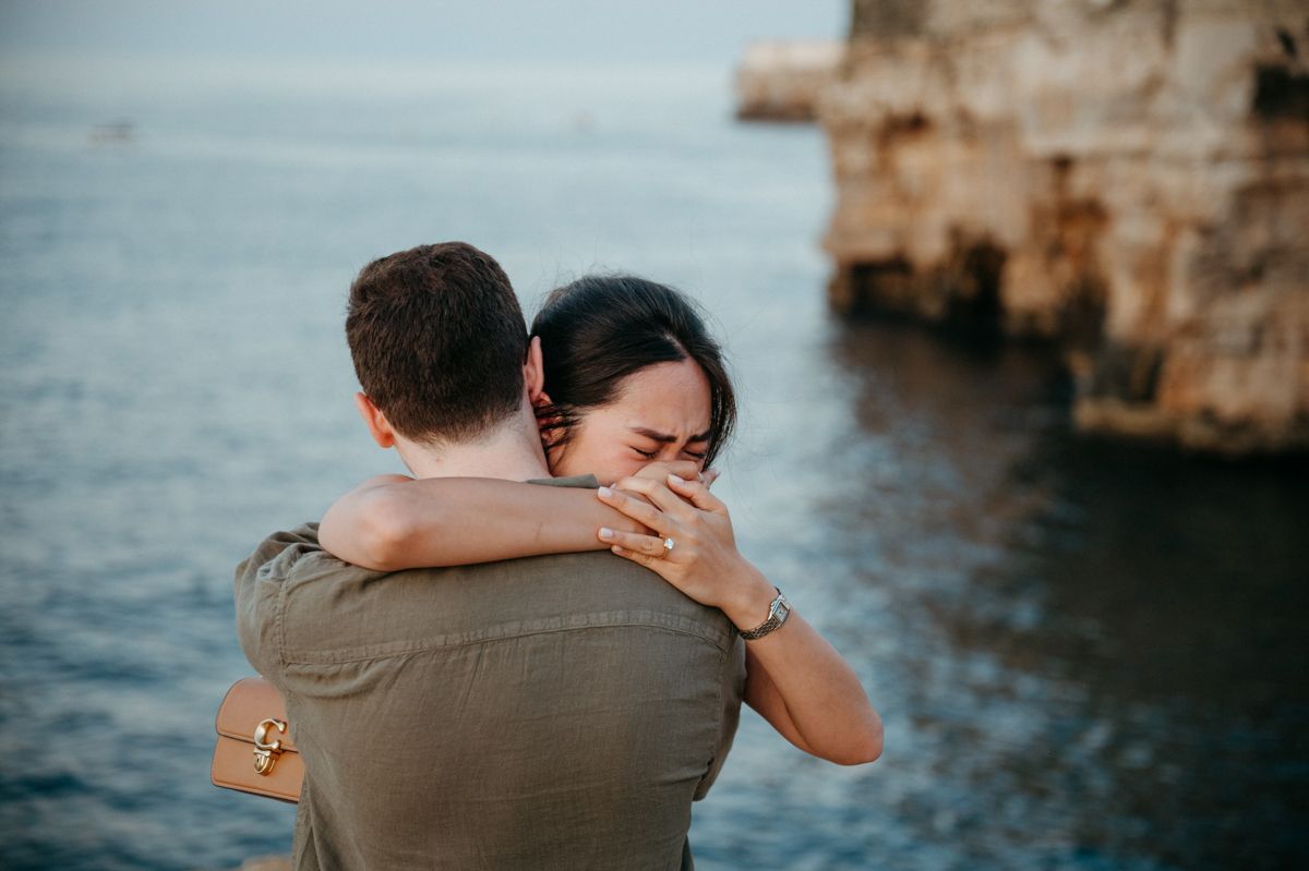 Two people embrace emotionally near rocky cliffs overlooking a blue ocean during golden hour.