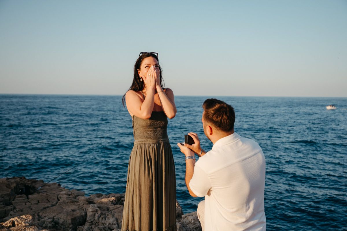 A surprise proposal moment unfolds on a rocky ocean coastline at sunset as waves crash in the background.