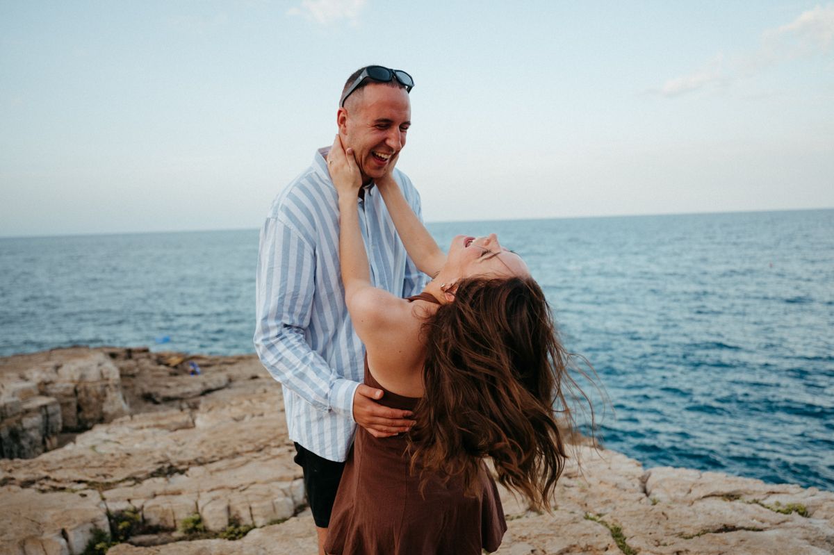 Romantic couple embracing on rocky coastal cliffs with ocean waves crashing in the background at sunset.