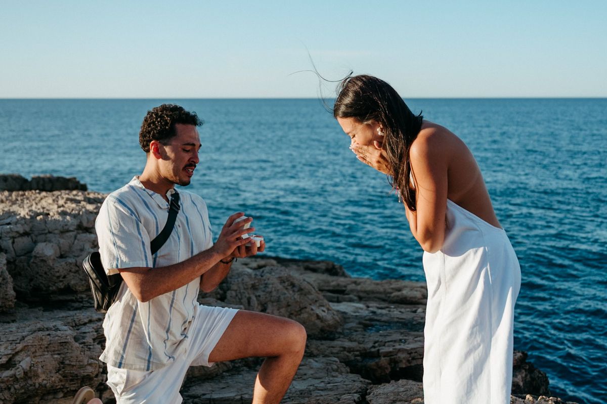 A couple enjoys a romantic seaside moment together on rocky coastal cliffs overlooking blue ocean waters.