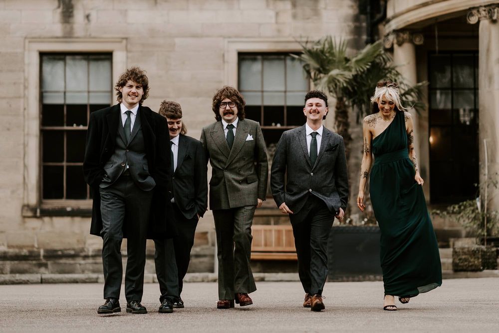 Wedding party members walk together on a paved pathway while wearing formal black suits and dresses.