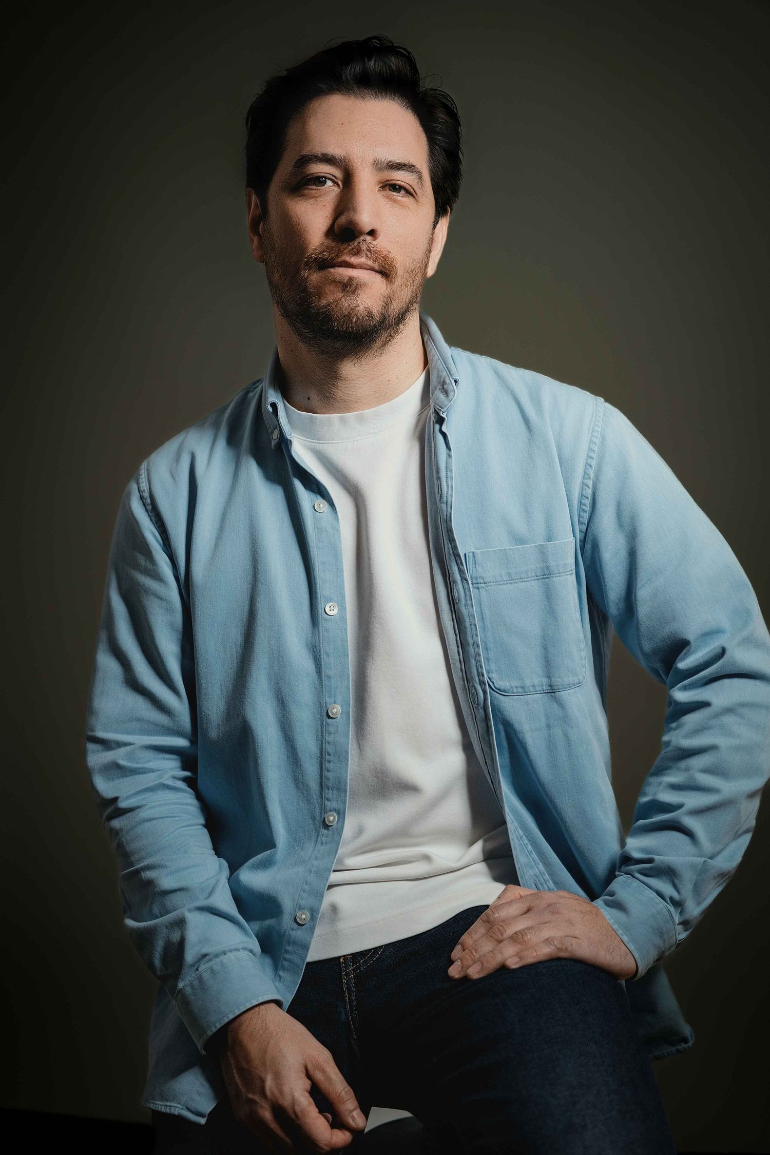 Studio portrait of someone in a light blue button-down shirt over white t-shirt against dark background.