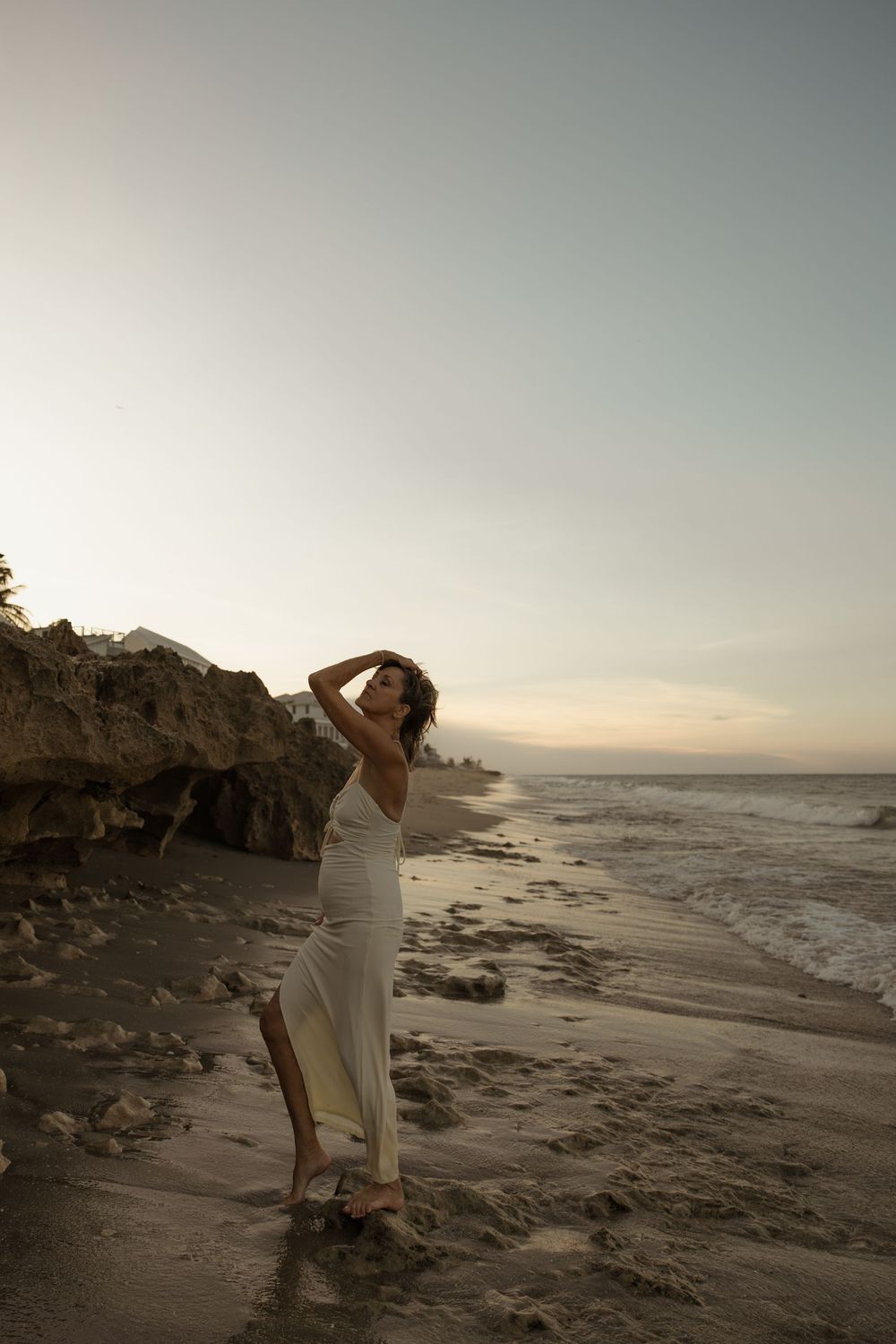A person in white stands on a rocky beach at sunset with ocean waves crashing against the rugged coastline.