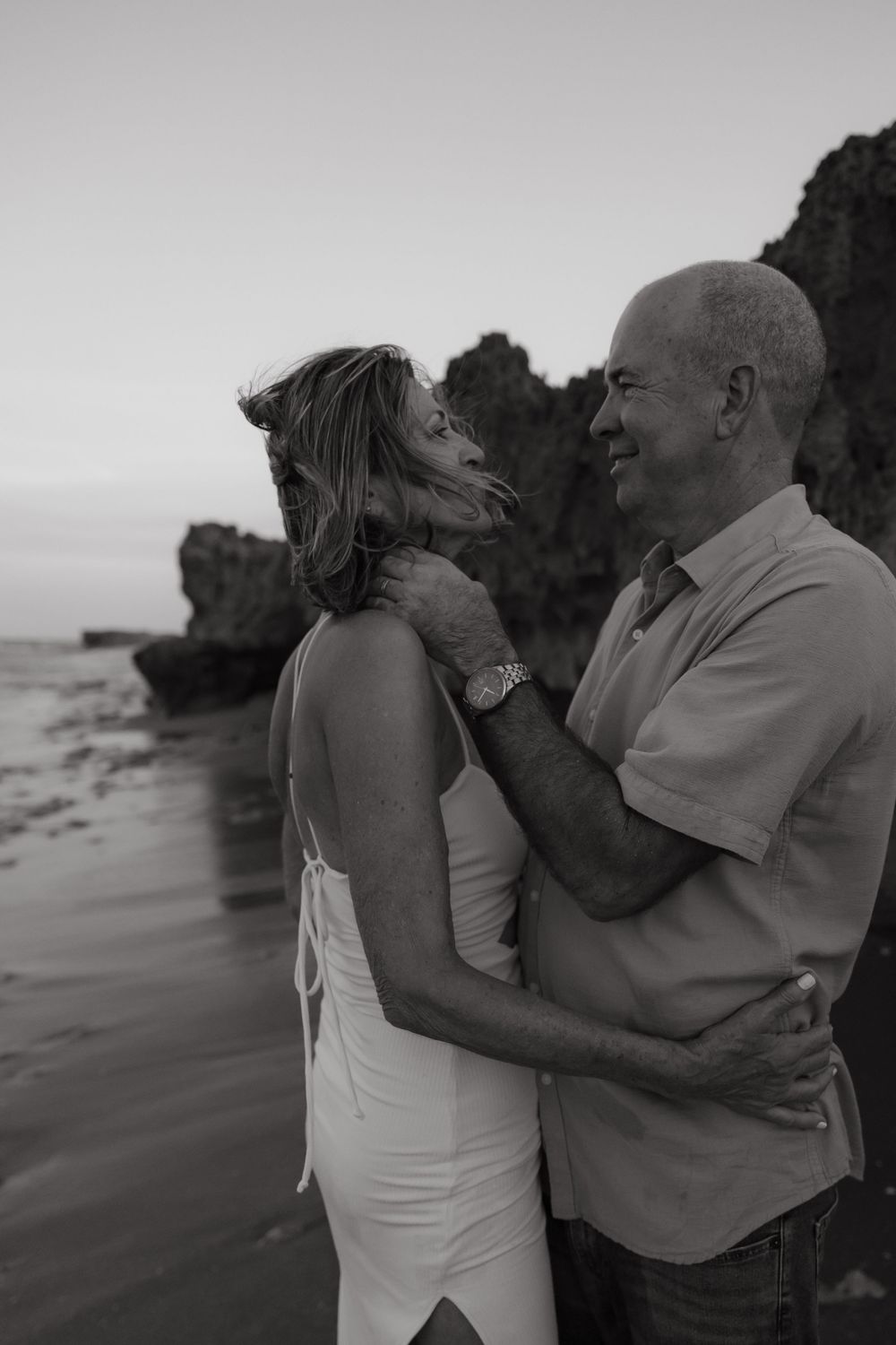 A romantic black and white photo of a couple embracing on a rocky beach at sunset.