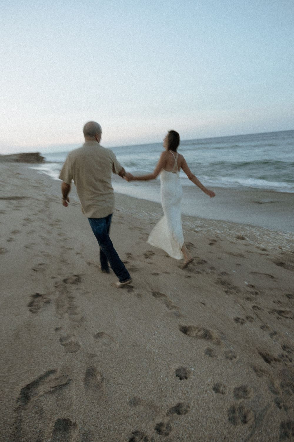 A couple in casual attire runs playfully along a sandy beach at sunset with ocean waves in the background.