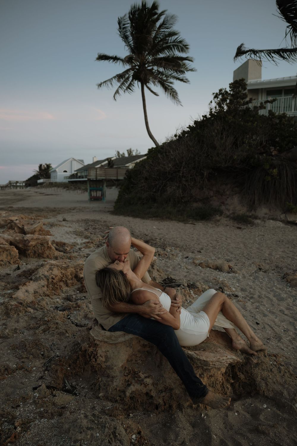 Couple relaxing on rocky beach at sunset with palm tree and buildings silhouetted against dusky sky.