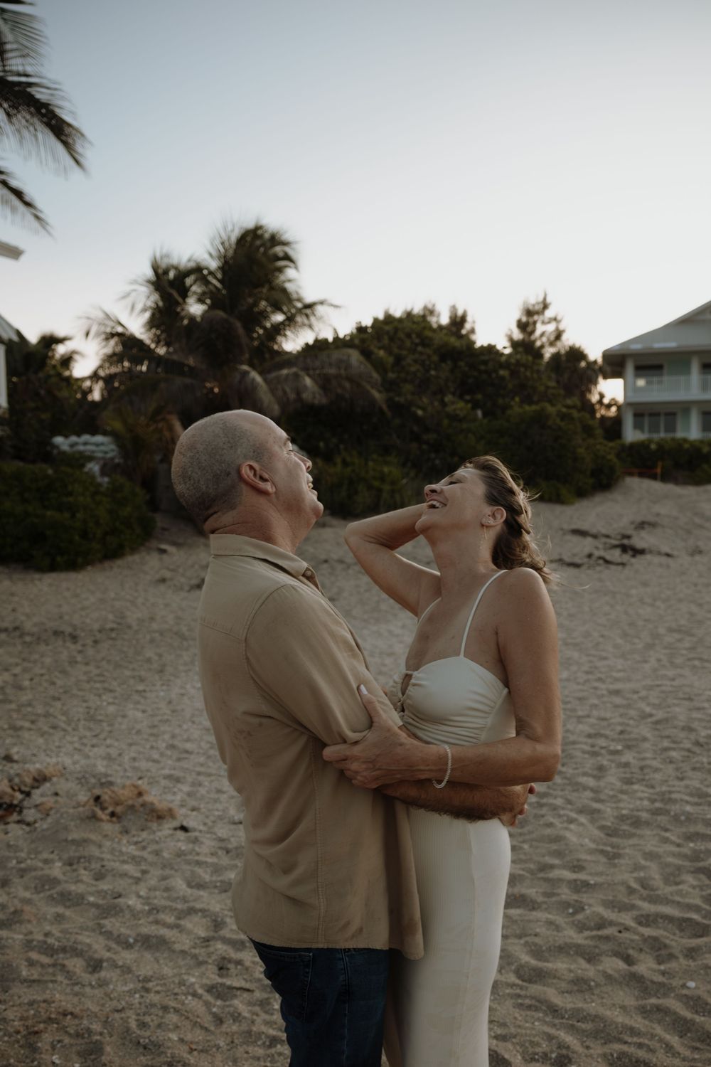 A couple shares a joyful moment together on a sandy beach at sunset with palm trees in the background.