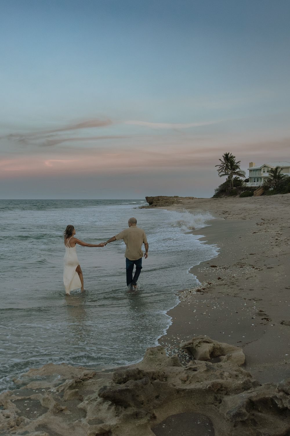 A couple walks hand in hand along a beach at sunset with a lighthouse and palm trees in the distance.