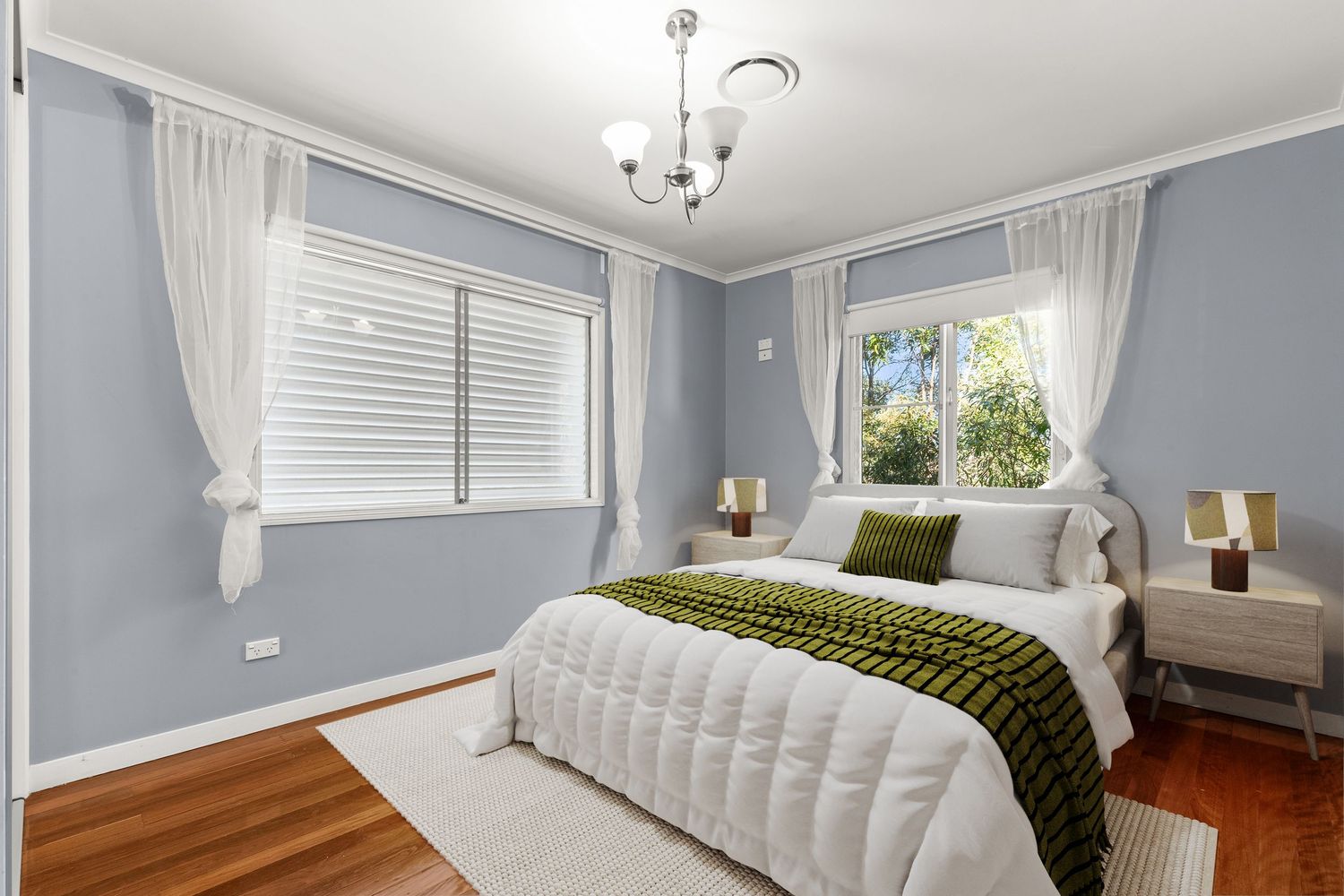 Elegant bedroom with blue-grey walls, hardwood floors, white bedding, plantation shutters, and a classic chandelier.