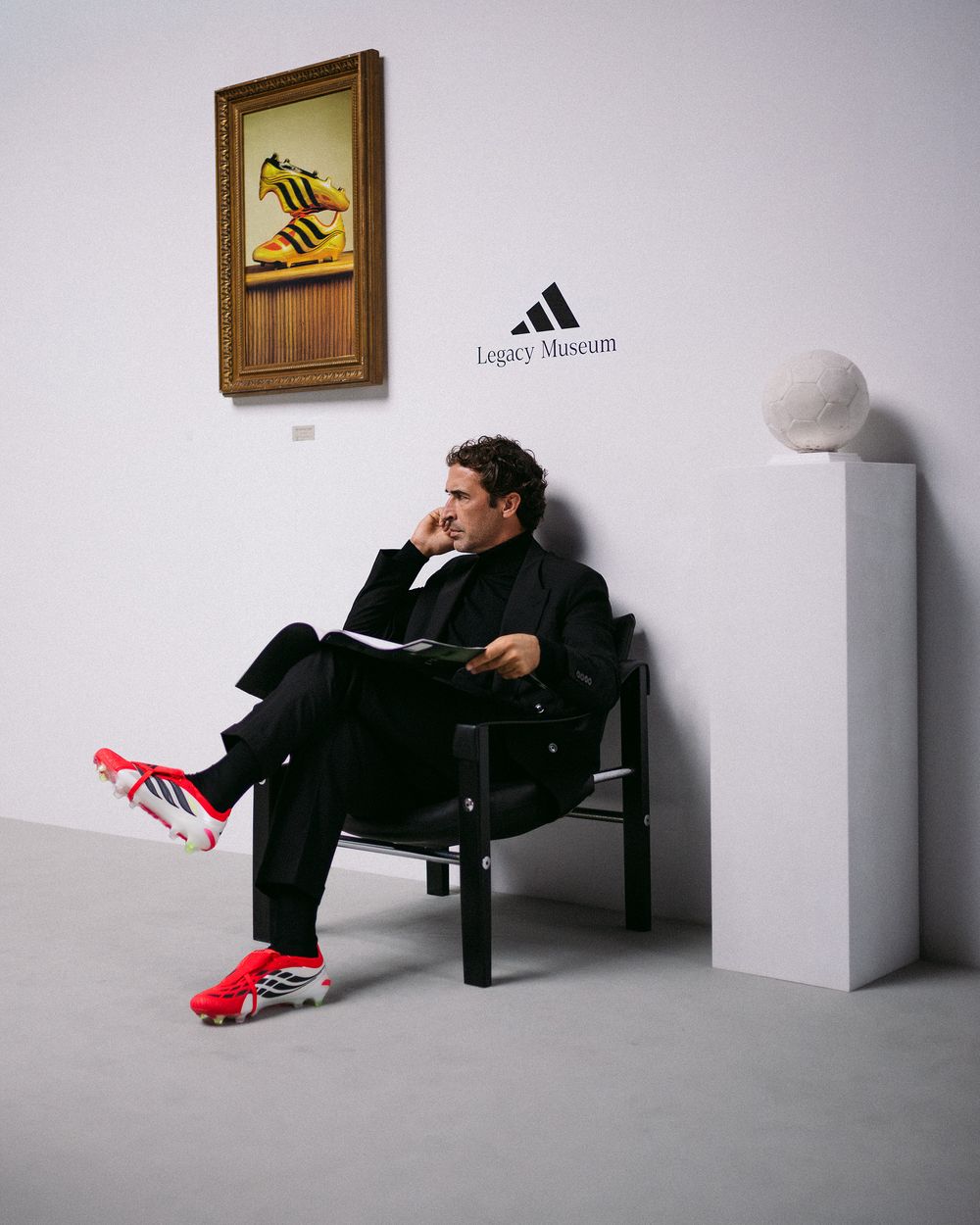 A person in black clothing lies casually on a white display pedestal next to an Adidas logo in a minimalist gallery setting.