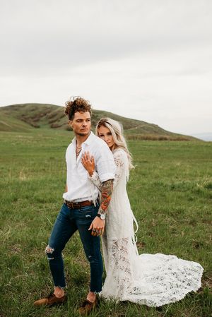 A couple in white wedding attire poses together on a green hillside with mountains in the background.