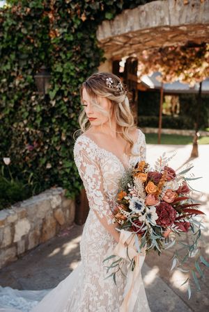 Bride in lace sleeve wedding dress holds vibrant autumn bouquet with burgundy and orange flowers against stone wall.