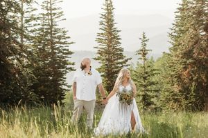 A couple in wedding attire holds hands while walking through a scenic mountain meadow surrounded by evergreen trees.
