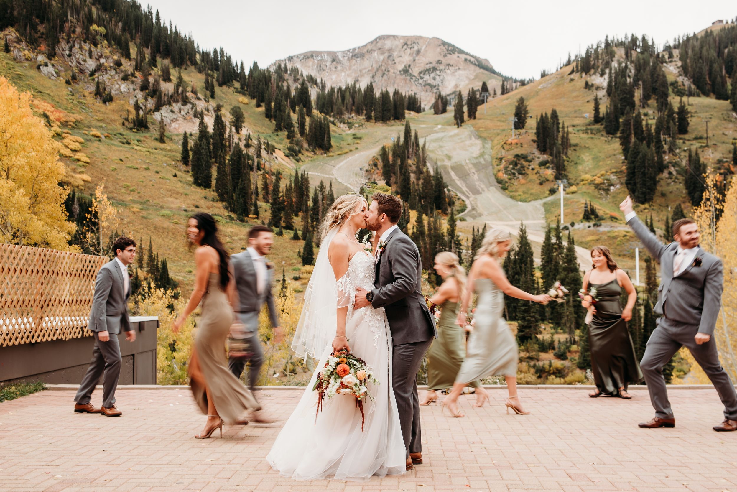 Wedding party dancing outdoors in a scenic mountain valley with evergreen trees and autumn colors in the background.