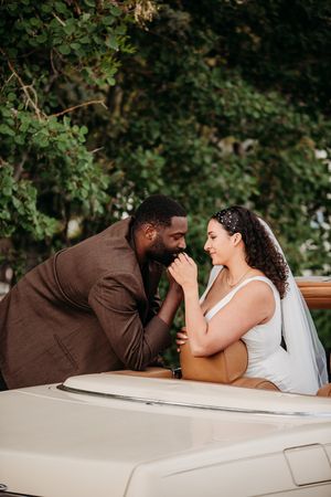 A couple shares a joyful moment while leaning on a classic car during their wedding photoshoot outdoors.