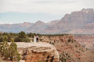 Two people stand on a scenic overlook with dramatic red cliffs and mountains of Zion National Park in the background.