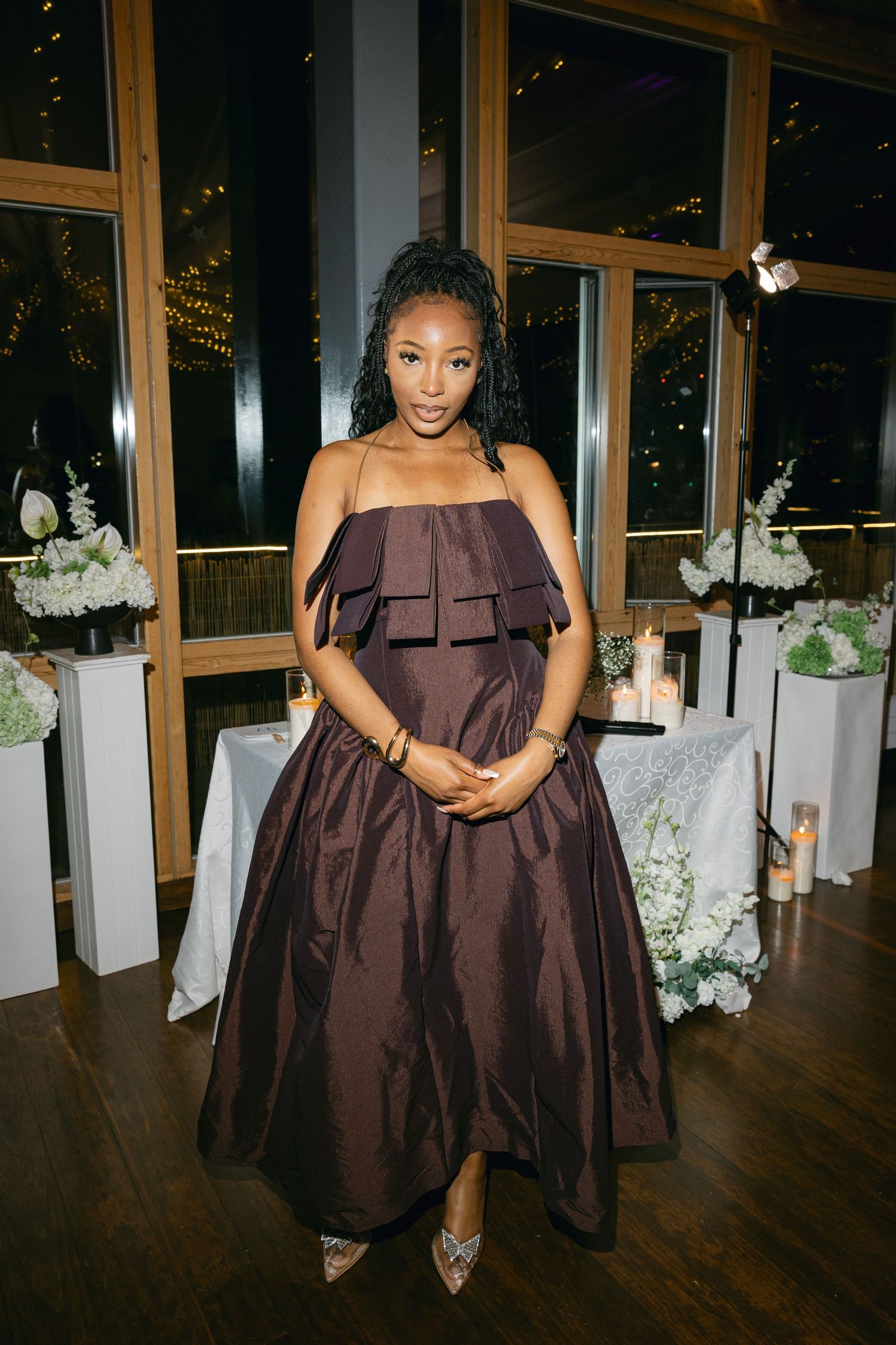 A woman in a chocolate brown off-shoulder gown stands in an elegant event space with white-draped tables.