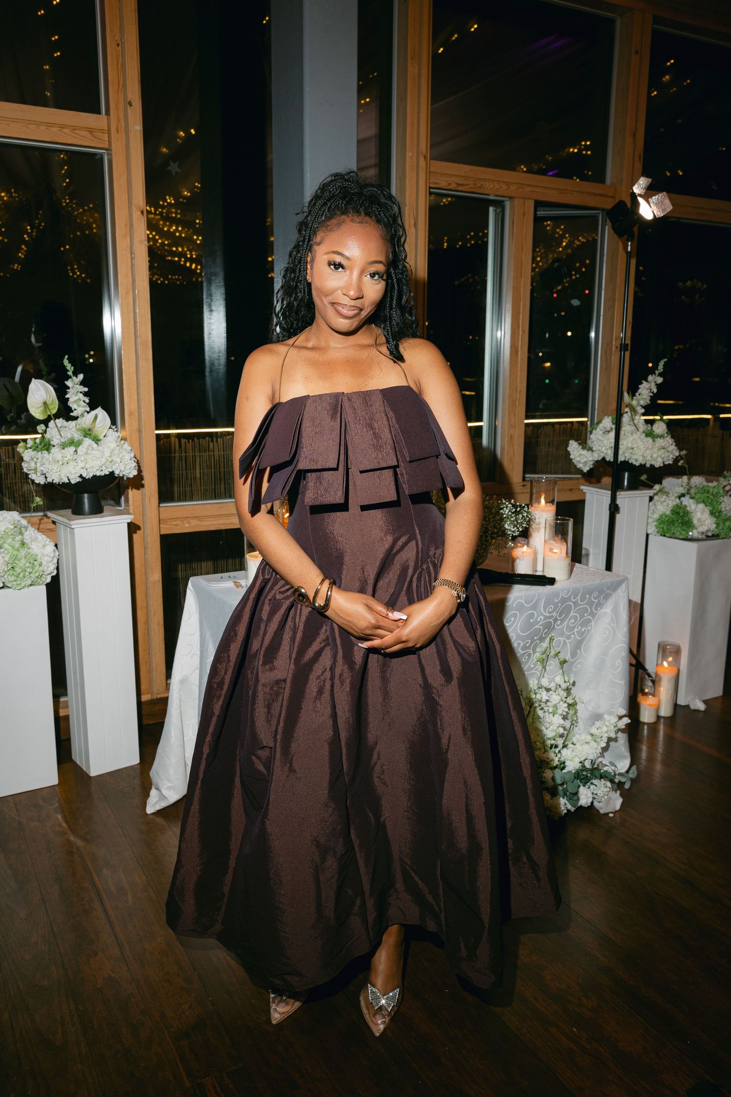 Wedding party member in a brown formal dress posing in front of windows at night.