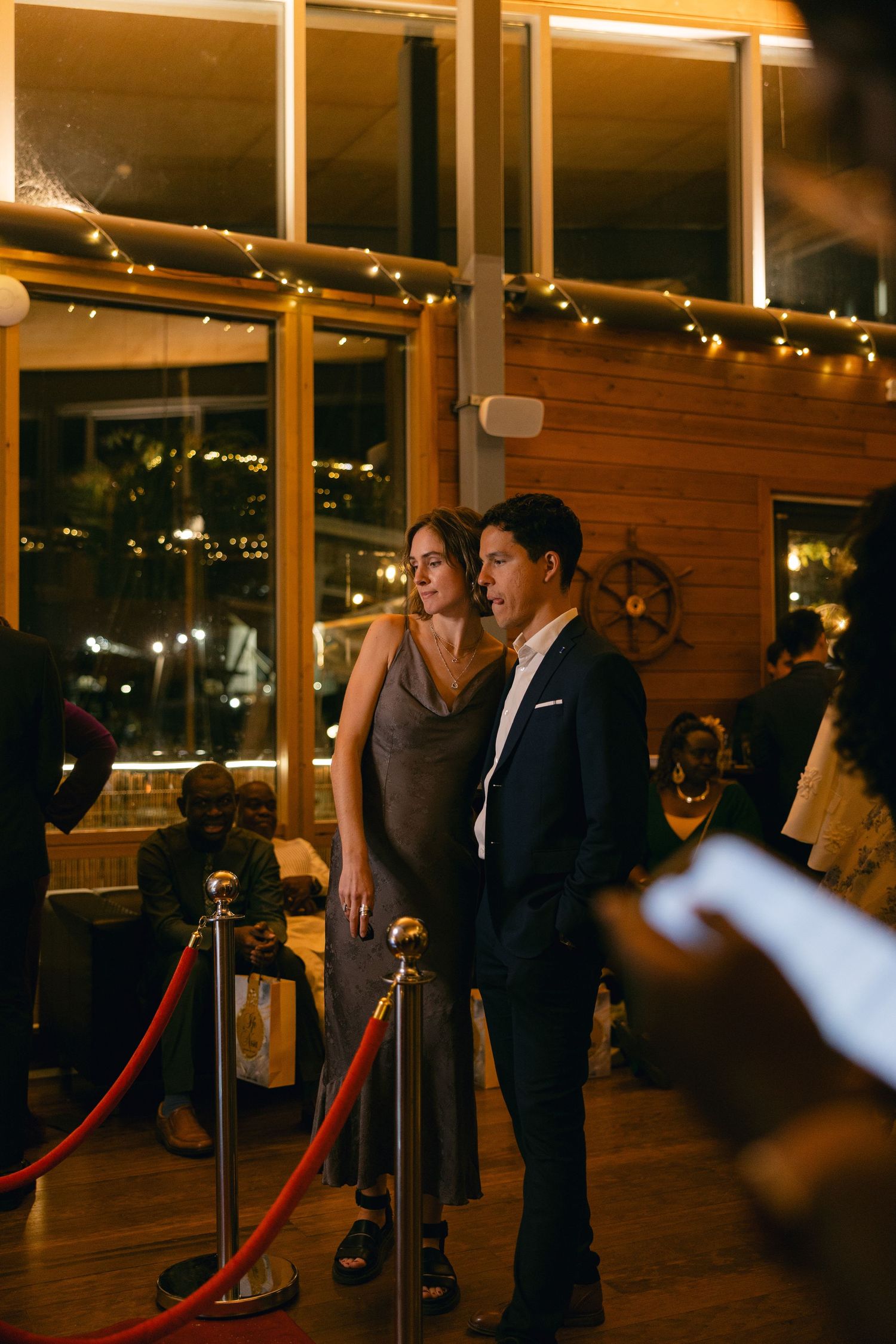 A couple poses for photos in a dimly lit venue with string lights and red velvet ropes creating an elegant atmosphere.